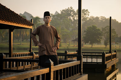 Portrait of man wearing traditional clothing while standing on built structure against lake during sunset
