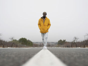 Ground level full body of african american male with hands in pockets standing on asphalt roadway among leafless trees and snow