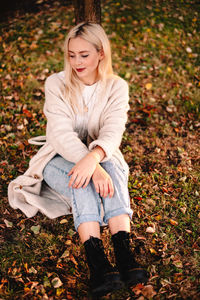 Happy young woman sitting on grassy field covered with fallen leaves