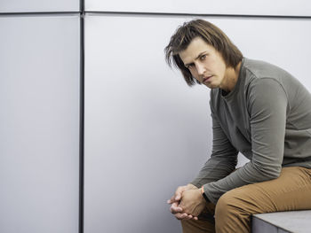 Millennial man with long hair and tense look sits near grey wall. serious man is staring in camera. 