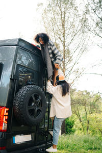 Side view of young african american woman helping friend climbing up camper van parked in green forest during summer adventure in countryside