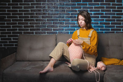 Full length of woman knitting while sitting on sofa at home