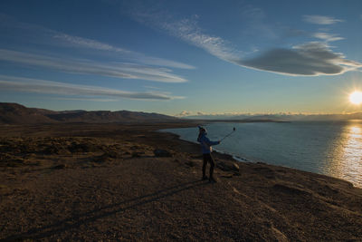 Woman taking selfie from monopod at lakeshore against sky during sunset