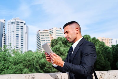 Side view of focused young hispanic male executive manager in classy suit with bag reading information on tablet while standing near railing on urban terrace