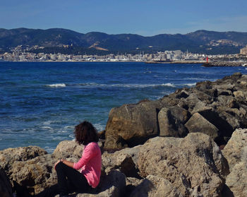 Rear view of woman sitting on rock by sea against sky