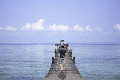 Rear view of man with crutches walking on pier over sea against sky