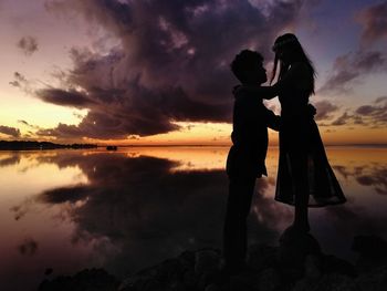 Silhouette couple standing at beach against cloudy sky during sunset