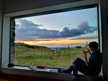 Woman sitting by window against sky during sunset