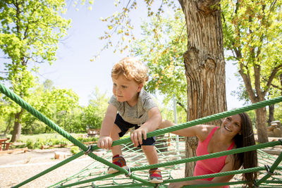 Smiling mother looking at son climbing on ropes in playground