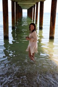 Portrait of smiling woman standing below pier amidst sea