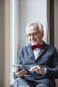 Smiling senior man sitting in windowsill using tablet