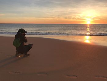 Man sitting on beach against sky during sunset