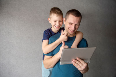 A cute boy hugs his dad from behind, talking on a video link in a tablet, showing a class