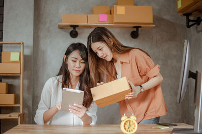 Woman holding box discussing with coworker using digital tablet in home office