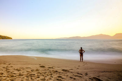 Rear view of woman standing at beach against sky during sunset