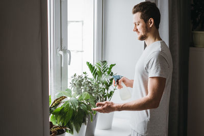 Side view of young man standing by window at home