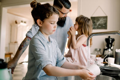 Girl washing mug at sink while father talking to daughter in kitchen