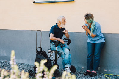 Smiling female nurse clapping while looking at senior man sitting on wheelchair against wall at back yard