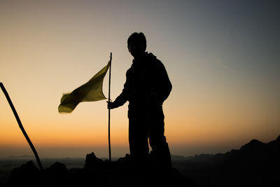 Silhouette hiker standing with flag on mountain against sky during sunset