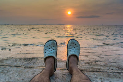 Low section of man relaxing on beach against sky during sunset