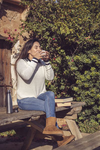 Latin woman having a cup of coffee sitting on a wooden bench in the garden