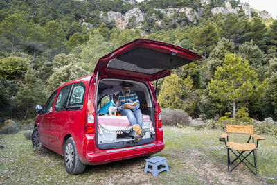 Full body of mature male reading interesting book while sitting in mini camper with french bulldog during road trip in countryside