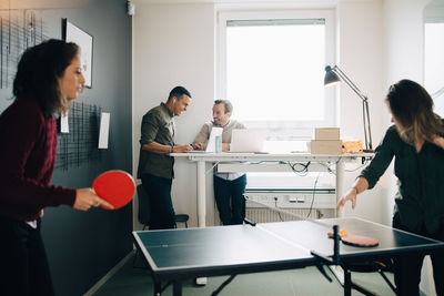 Female professionals playing table tennis while male colleagues using laptop at creative office