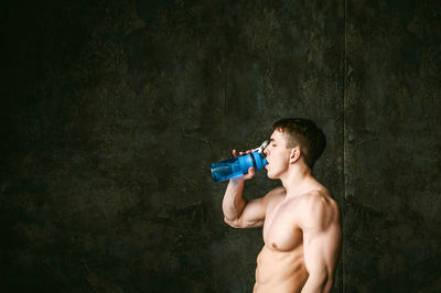 Shirtless man drinking while gesturing thumbs up by wall