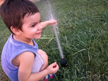 Cheerful boy playing with sprinkler while crouching on field