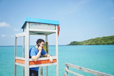 Man sitting on seat by sea against blue sky