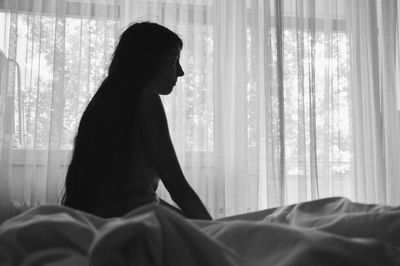 Silhouette woman looking away while sitting on bed against curtain at home