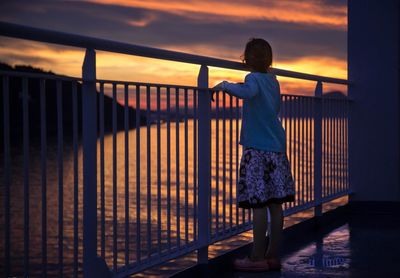 Rear view of boy standing on railing against sky during sunset