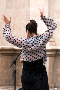 An averted woman in a dotted blouse raises her hands to sing or dance in the street