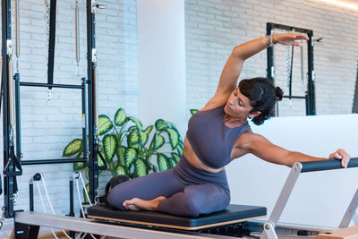 Slim adult female doing seated side bend and stretching body on modern reformer during pilates training in gym