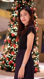 Portrait of mid adult woman standing against illuminated christmas tree