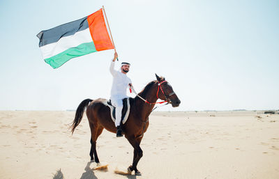 Full length of man with flag riding horse on desert against sky