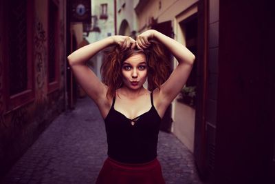 Portrait of young woman with hand in hair while standing on in aisle of buildings