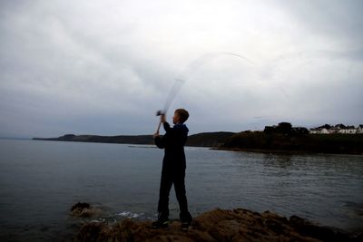 Man fishing on rock by sea against sky