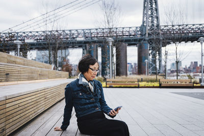 Senior woman using smart phone while sitting on seats against bridge in city