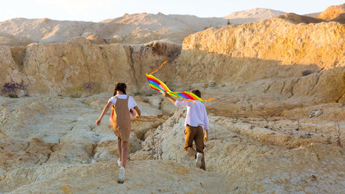A girl and boy are running around a sandy quarry with bright kite.