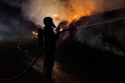 Rear view of man standing by illuminated fire against sky at night