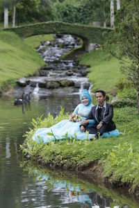 Portrait of bride and bridegroom sitting against waterfall at park