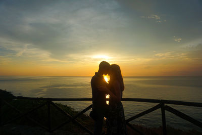 Side view of couple kissing while standing by sea against sky during sunset
