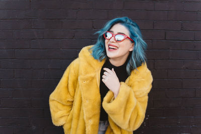 Smiling woman wearing coat and sunglasses while standing against brick wall