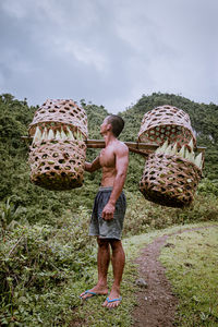 Full length of shirtless man standing in basket on field against sky