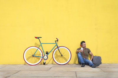 Man using phone while sitting with bicycle against yellow wall