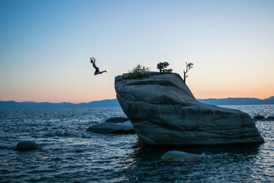 Man jumping off of rock by sea against clear sky