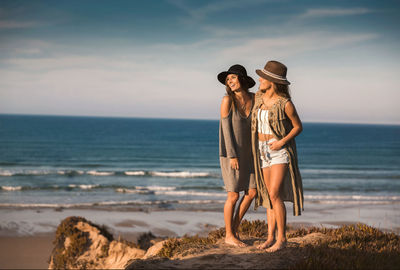 Female friends wearing hats while standing at beach against sky