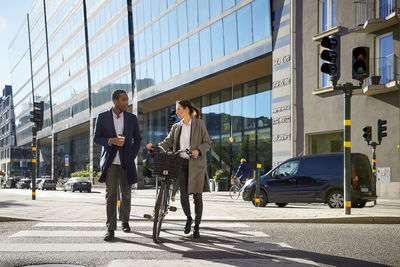 Full length of woman and man talking while walking with electric bicycle on crosswalk against building in city