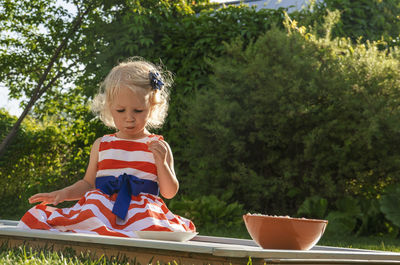 Pretty curly little girl seating in garden and eating marshmallows from the plate.